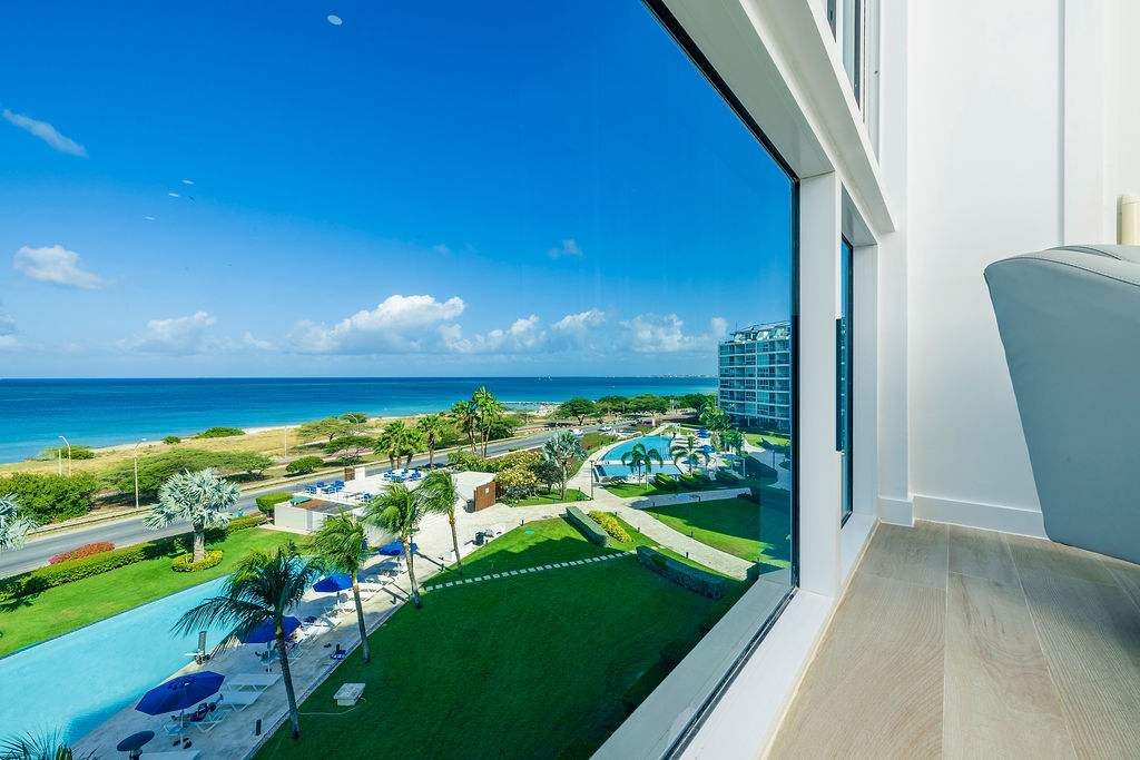 Stunning ocean views unfold through floor-to-ceiling windows, showcasing turquoise waters, tropical palms, and resort amenities below your elevated retreat.