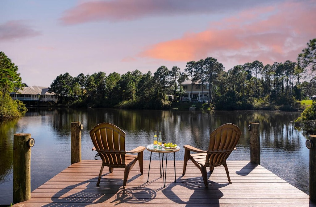 Waterfront dock at Serenity Lake with Adirondack chairs overlooking tranquil lake at sunset, surrounded by trees and nearby homes.
