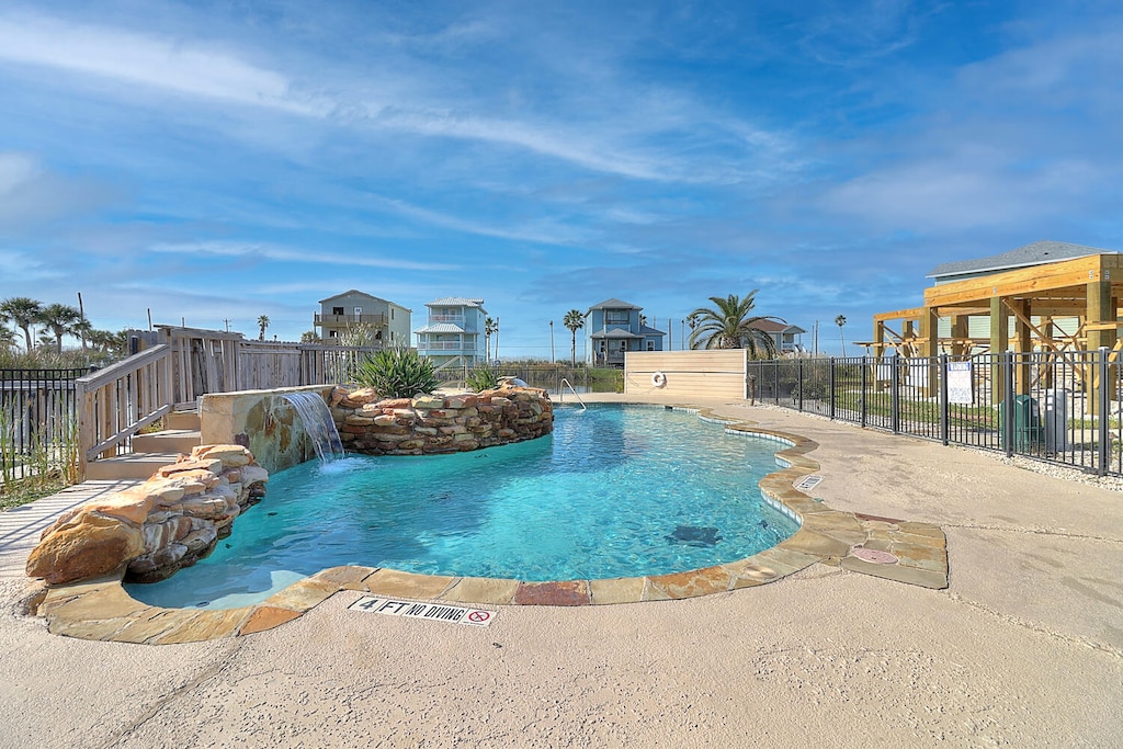 Resort-style pool with waterfall feature and coastal property views under sunny blue skies.