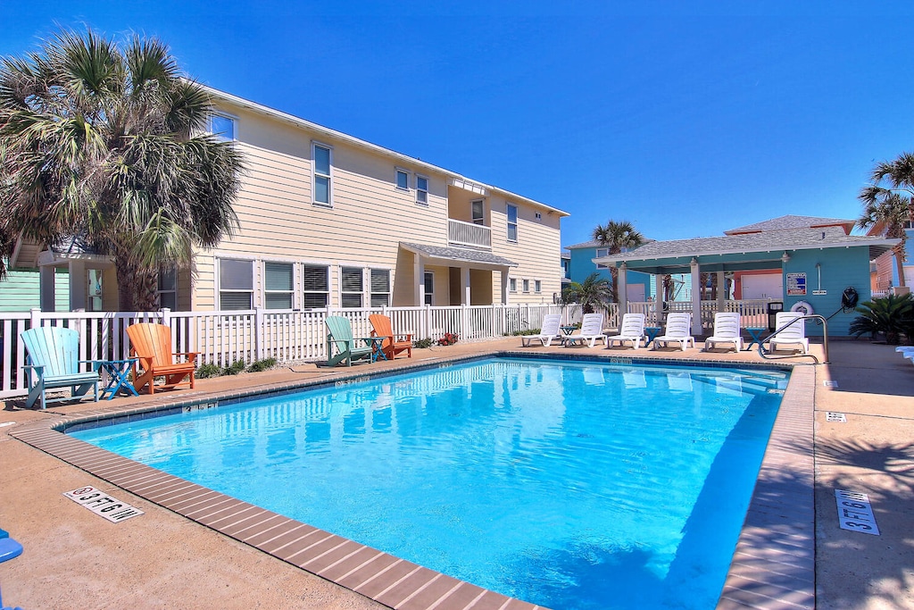 Coastal vacation rental complex featuring a sparkling blue swimming pool surrounded by colorful beach-style buildings and palm trees.