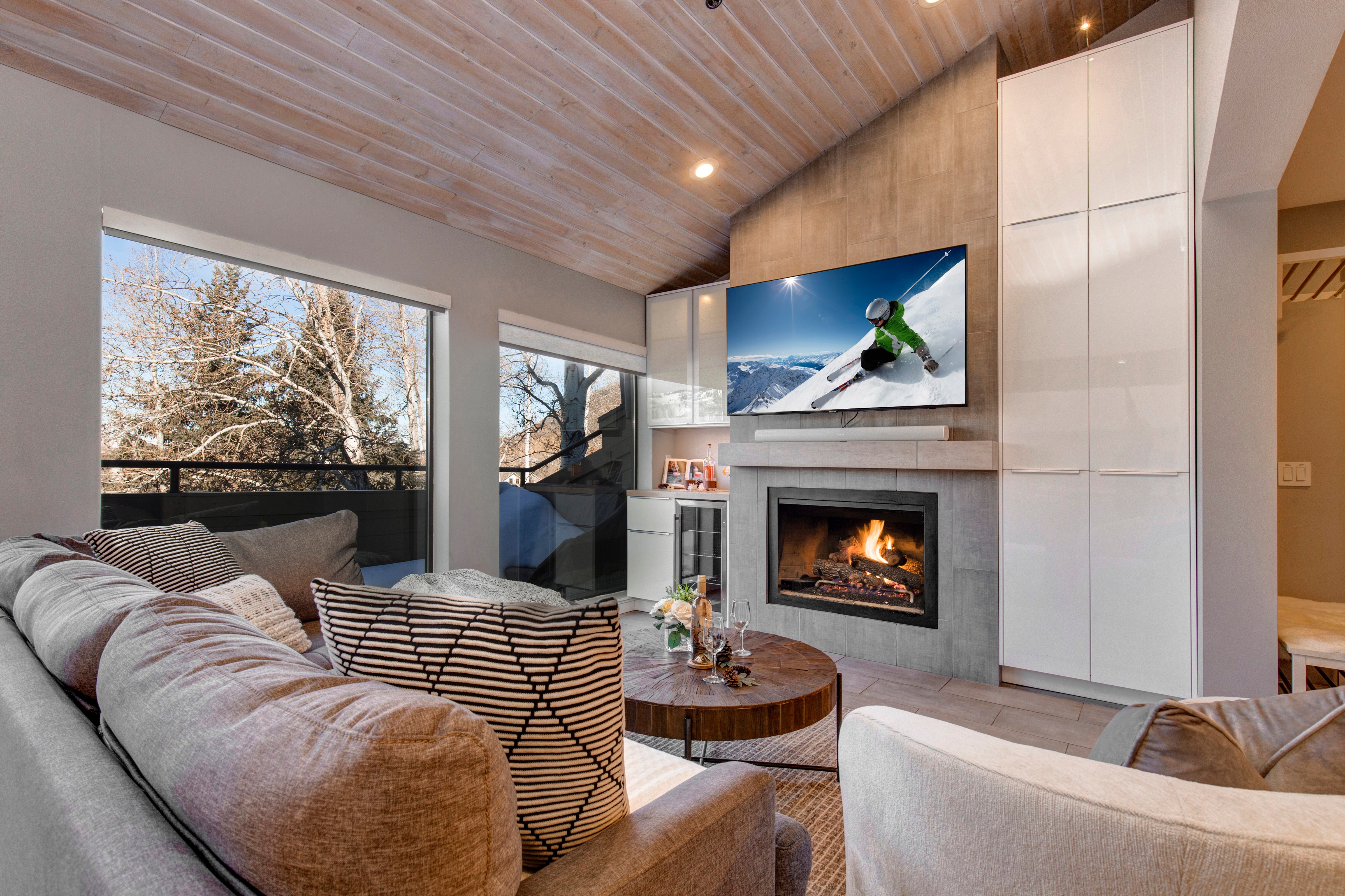 Living Room with plush sectional and arm chair, gas fireplace, 65" smart tv, wet bar with wine fridge,  built-in work station, and private deck access